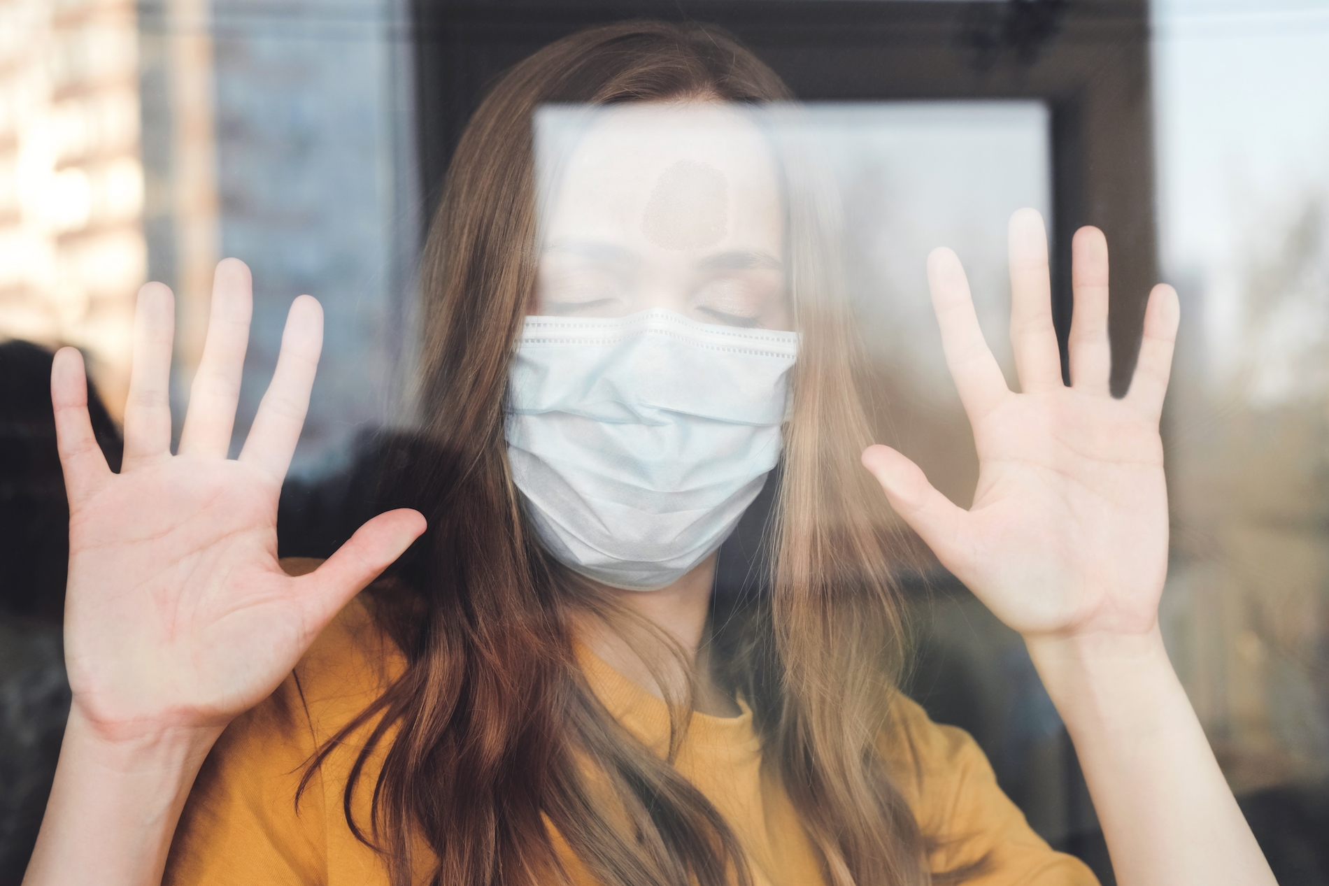 Sad girl in medical protective mask and yellow T-shirt standing at her house and looks out the window. Covid-19 coronavirus pandemic. Isolation at home. Protect yourself and your loved ones.