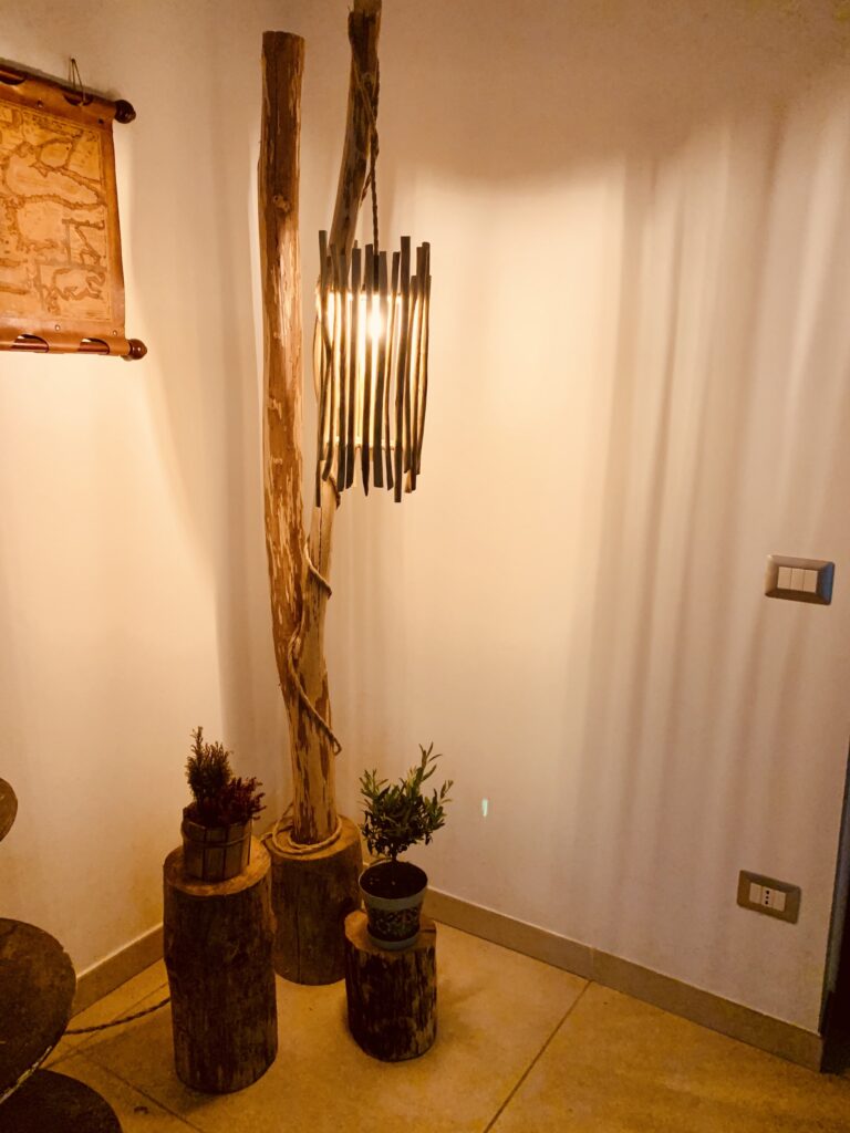 Una lampada da terra, scultorea e nobile nel suo aspetto grezzo, si erge come un antico albero che sussurra storie al vento. Intorno, sgabelli di legno massiccio, anch'essi testimoni della forza silenziosa della natura, e piccole piante verdi, un bonsai di olivo, simboli di vita e rinnovamento. La luce, filtrata da rametti disposti come le sbarre di un carillon, si diffonde in onde calde che accarezzano le pareti con ombre morbide e invitanti.