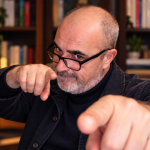 Carlo D’Angiò indica verso la fotocamera in uno studio con libreria sullo sfondo